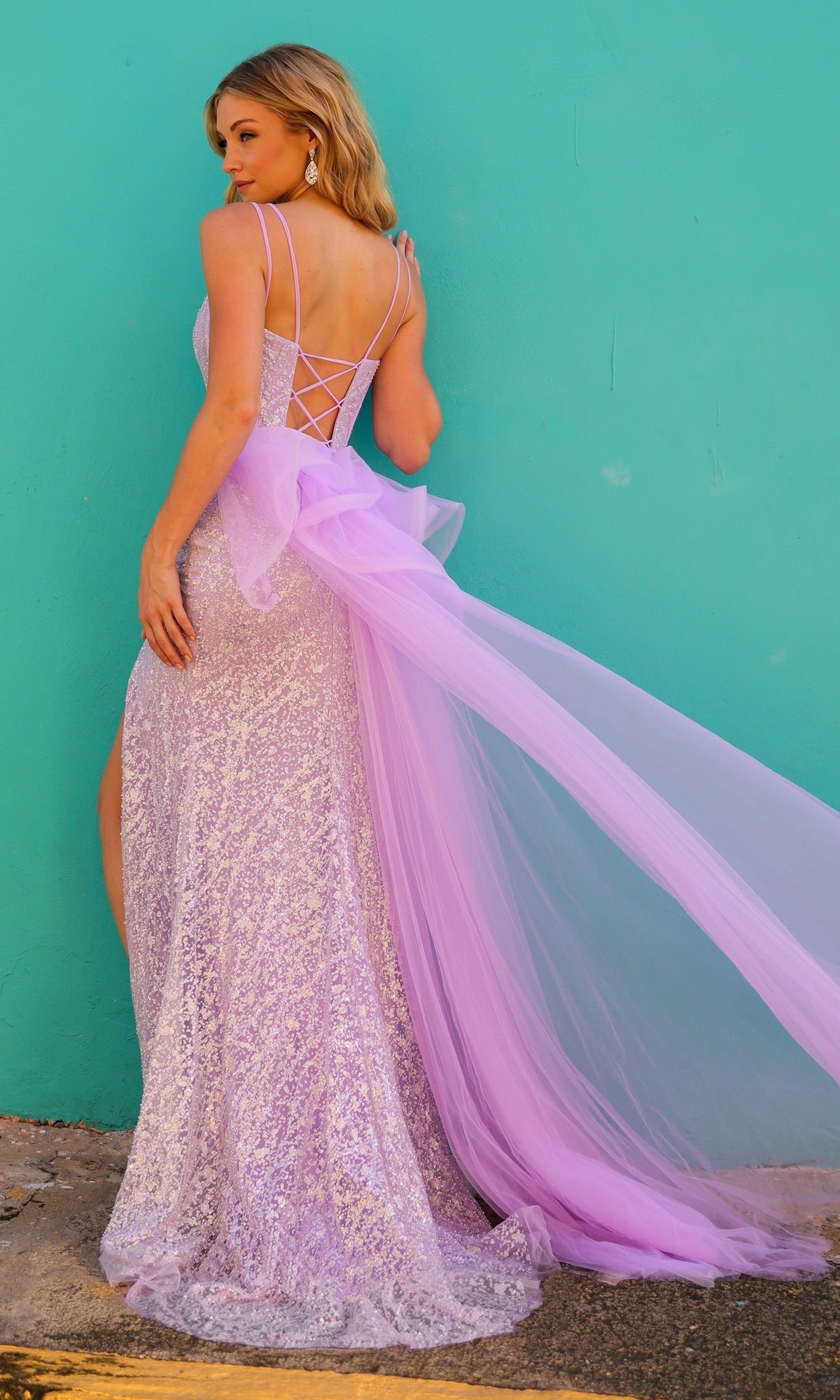 A woman wears the Glitter-Print Long Prom Dress with Overskirt Y1475, featuring a crisscross back and long tulle bow, standing against a turquoise wall and glancing over her shoulder.